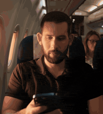 איילון יושב במושב חלון במטוס ומסתכל בטלפון, ואז עומד על סיפון של ספינה בסערה, עם שמיים אפורים וגלים מאחוריו, ומקליד בטלפון